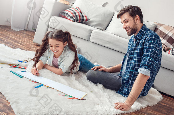 父亲和小女儿在家里的地板上，男人看着女儿在<strong>画册</strong>上微笑着画画