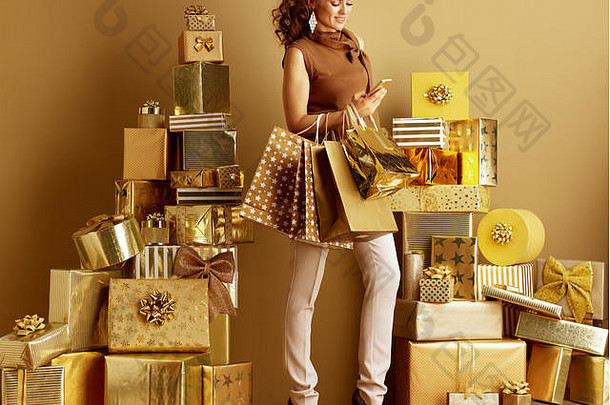 完整的长度肖像微笑年轻的购物者女人黄金米色裤子棕色（的）上衣购物袋写作短信桩金礼物