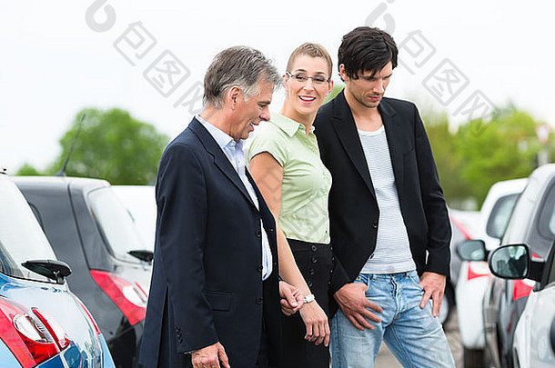 成熟的汽车经销商和站在车前经销商停车场的年轻夫妇