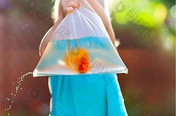 一个穿着蓝色衣服的小女孩站在街上，手里拿着一个透明的塑料袋，里面装着水和金鱼，水从小孔里滴了下来