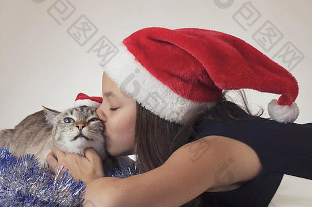 美丽的少女和她戴着圣诞老人帽子的猫在白色背景上快乐地接吻