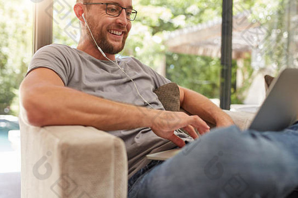 微笑的成熟男子在家里使用笔记本电脑，他坐在沙发上戴着耳机。