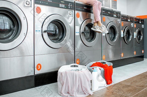 一名妇女坐在洗衣机上，拿着装满衣服的篮子在自助洗衣店等待洗衣。无面裁剪图像