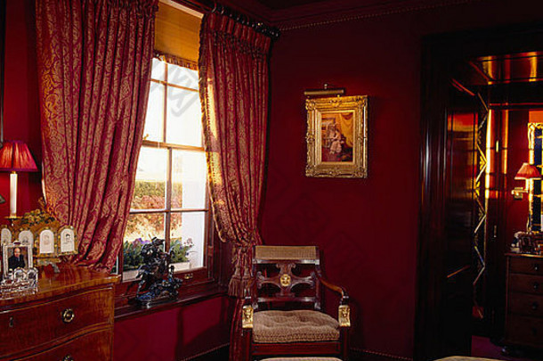 深红色卧室的椅子和脚凳上方有镀金框架的发光图片，带有图案红色窗帘和米色百叶窗