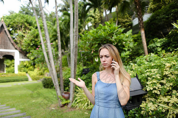 一个穿着牛仔裤和太阳裙的年轻女子在树旁的花园里用智能手机聊天。