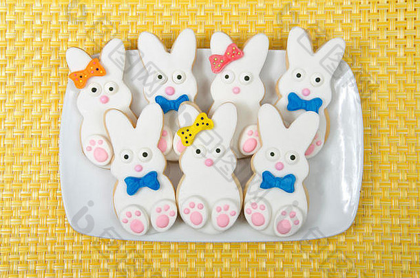 复活节兔子糖饼干放在一个盘子里，盘子的背景是黄色的垫子。棉花糖软糖覆盖的饼干，有糖果脚、眼睛、鼻子、尾巴和尾巴