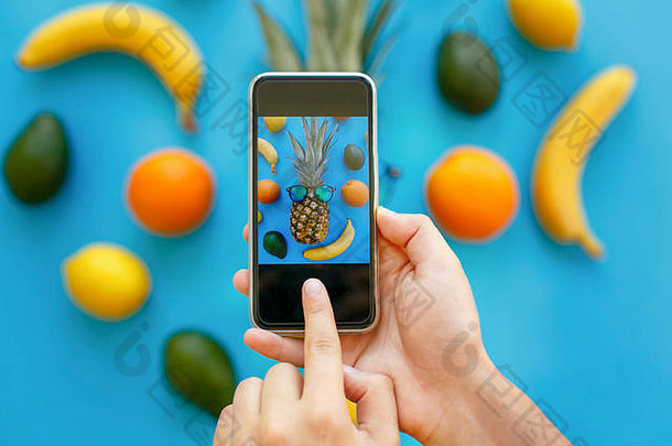 手拿着手机，在蓝色的纸上拍着戴着太阳镜的菠萝、香蕉、橘子和鳄梨的照片，时尚的平面。时尚食品摄影