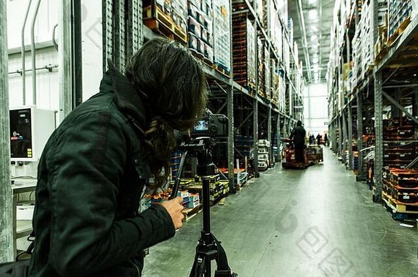 一名摄像师站在仓库内一个在三脚架上的专业单反相机后面，为公司拍摄宣传媒体，右边是复印空间。
