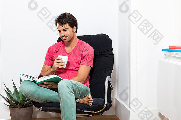 一个穿着休闲服的年轻人坐在黑色的椅子上看书，手里拿着一杯饮料。