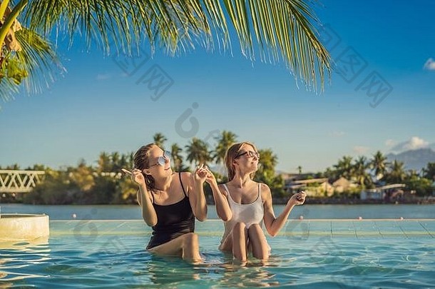 暑假期间，两位美丽的年轻女子坐在度假胜地游泳池的池边。热带国家的聚会、旅游