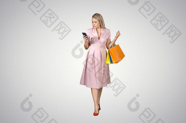 带着购物袋的购物妇女在梯度背景上行走时在智能手机上发短信。