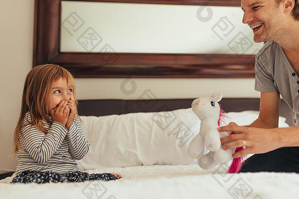 父亲拿着玩具在家里和女儿玩。快乐的小女孩手捂着嘴笑着，和坐在床上的父亲玩耍。