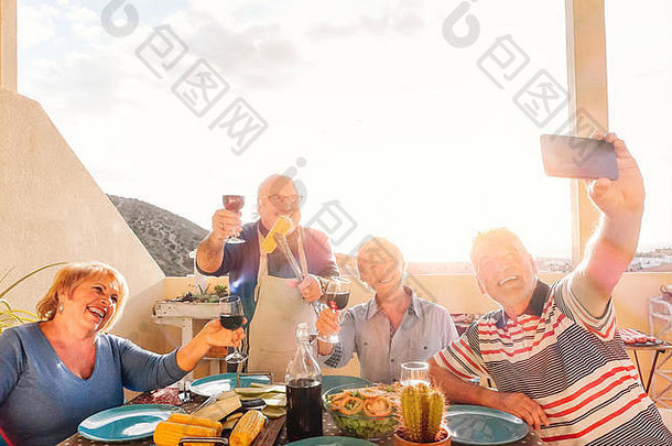 快乐的老年人用手机自拍，在屋顶烤肉，退休的人享受着吃红酒的乐趣