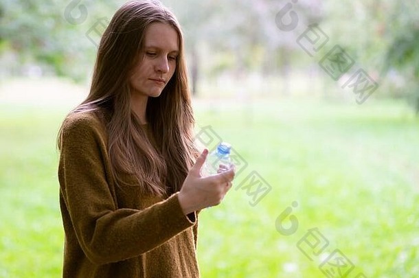 秋冬时节，在公园的街道上，年轻漂亮的女孩正在用塑料瓶喝水。一个留着漂亮又长又厚的黑头发的女人