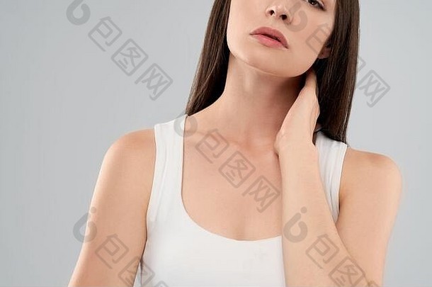 一名穿着白衬衫的不知名白人妇女在灰色隔离背景上摆姿势，因疼痛用手触摸脖子。前茬黑发用嘴呼吸。