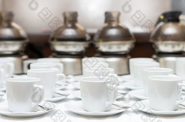 桌上和咖啡机背景上有许多排白色咖啡杯，带有茶碟和茶匙