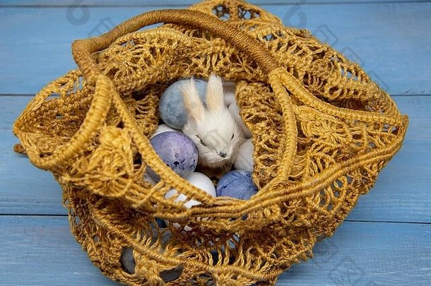 玩具兔子和复活节彩蛋装在柳条袋中，蓝色木质背景，采用环保材料制成。