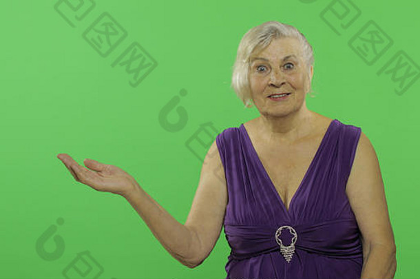 一位上了年纪的妇女用张开的手掌指着呈现的东西，微笑着。穿着紫色连衣裙的漂亮老奶奶。放置您的徽标或文字。色度键。绿色屏幕<strong>背景</strong>