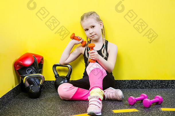 主题：运动与健康儿童。一个留着辫子的有趣的白人小女孩，坐在健身房的地板上休息。运动员哑铃器材