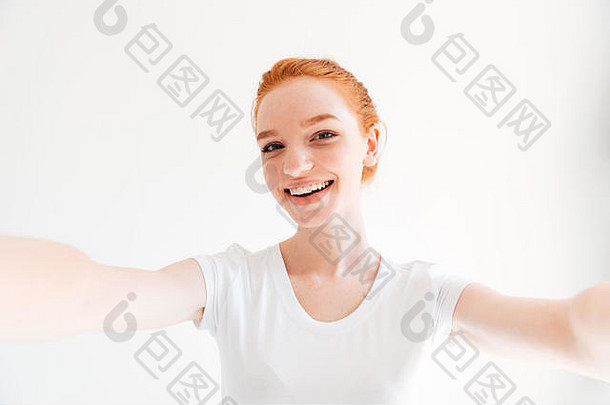 穿着t恤衫的快乐姜女自拍，在白色背景下看着相机