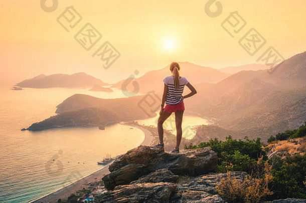 年轻女子站在岩石顶上，看着海滩和群山，看着夏日五彩缤纷的夕阳。风景与女孩，海，山
