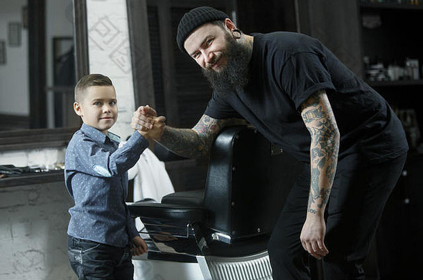 <strong>儿童</strong>理发师和小男孩在剪发后的深色背景下。大师的手上有“剃须”这个字的纹身