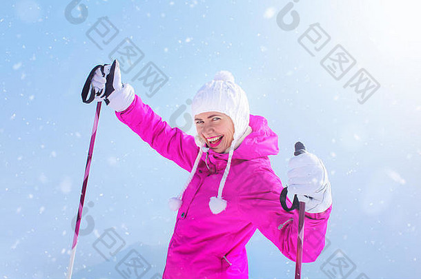 穿着粉色滑雪夹克、戴着手套拿着滑雪杆的快乐的年轻女子，在阳光和雪的映衬下微笑着。