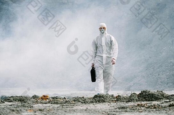 实验室技术员戴着面具，穿着化学防护服，拿着工具箱在干燥的地面上穿过有毒烟雾