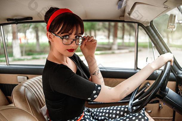 身着黑色复古连衣裙、戴复古眼镜的白人美丽年轻女孩的肖像，在一辆复古汽车的轮子后面摆姿势