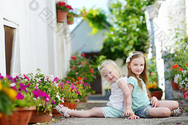 两个可爱的小姐妹坐在花盆之间，在温暖阳光明媚的夏日，在意大利小镇