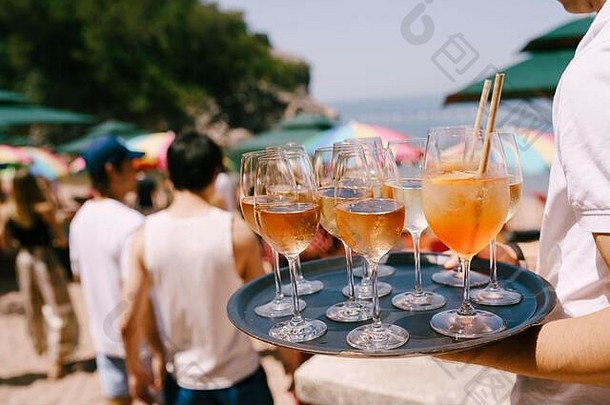 在海滩上的一个聚会上，一个手里拿着托盘的服务员端着鸡尾酒和饮料——一杯装在大杯里的橙色鸡尾酒，里面放着一管开胃酒和一杯威士忌