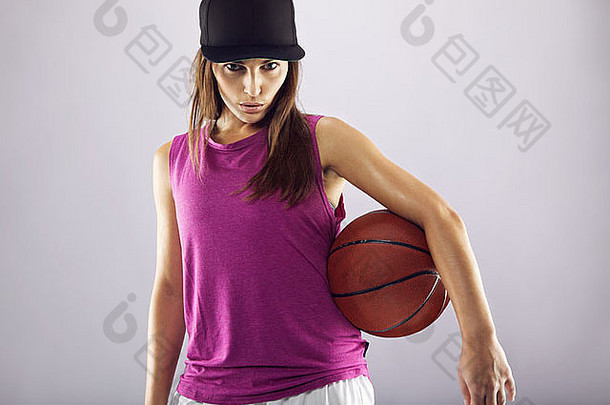 在灰色背景下持球的女篮球运动员。拿着篮球的年轻女子自信地看着摄像机