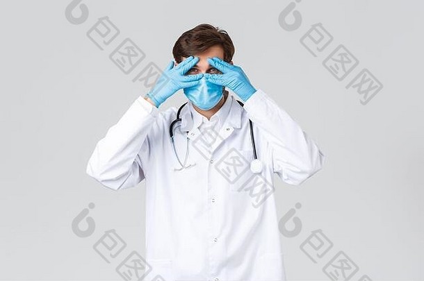 医院、医疗工作者2019冠状病毒疾病治疗理念。英俊的医生戴着医用口罩，磨砂膏和手套，透过手指看，偷看眼睛
