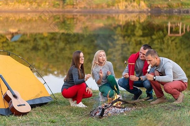 旅行，旅游，远足，野餐和人的概念-一群快乐的朋友在湖附近的篝火上炸香肠
