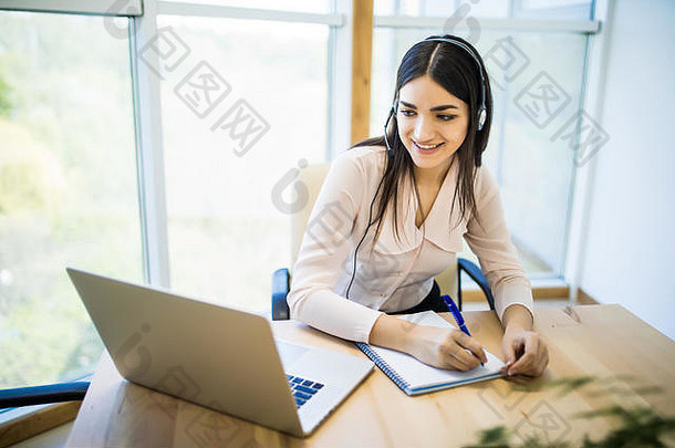 在办公室里，一位快乐的年轻女士坐在笔记本电脑旁使用耳机工作