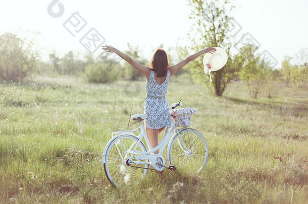 这位年轻女子正背着身子站着。她靠在自行车上，举起双手。