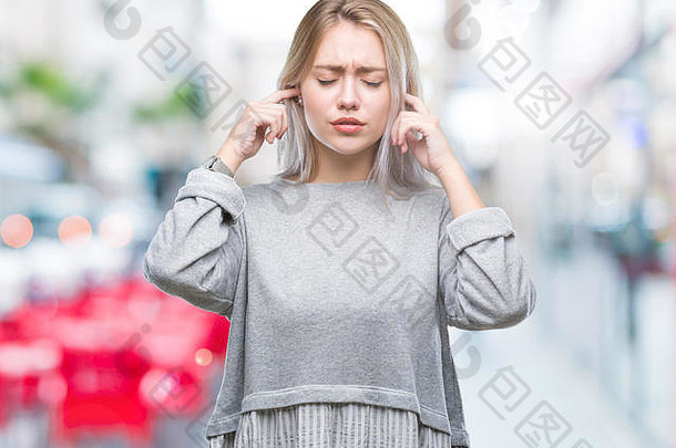 孤立背景下，年轻的金发女郎用手指捂住耳朵，对吵闹的音乐声表现出恼怒的表情。聋哑人的概念。