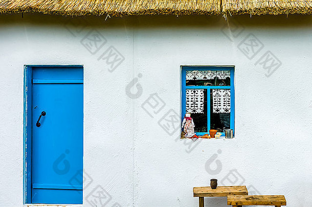 乡村农舍的白色墙壁，有一扇蓝色的门和一扇窗户。窗台上有布娃娃、花盆和一个锡杯
