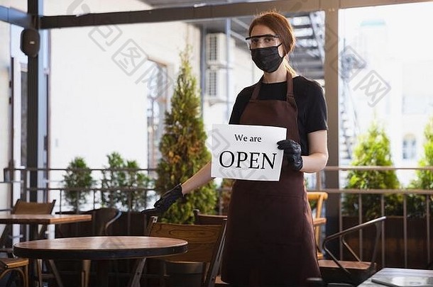 在冠状病毒大流行期间，这位女服务员戴着医用口罩、手套在餐厅工作。代表服务和安全的新常态。用文字支撑着木板，我们是开放的。保持距离。