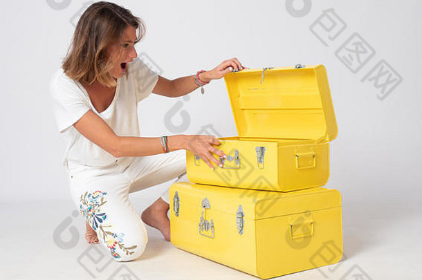 一个穿着白色衣服的金发女孩在摄影棚里摆出姿势，旁边放着两个黄色的金属手提箱。
