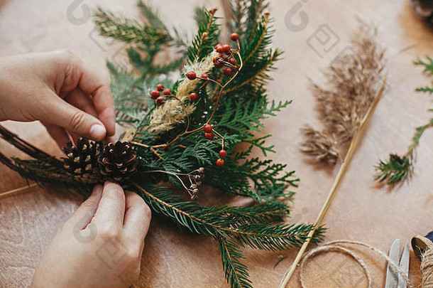 制作质朴的圣诞花环。手拿松果、浆果、冷杉枝、线、剪刀放在木桌上。正宗的乡村花环。圣诞节