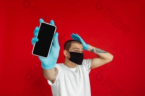 一个戴着防护面罩和医用手套的白人年轻人在红色工作室的背景下展示了手机的空白屏幕。人类情感的概念，销售，冠状病毒锁<strong>定</strong>，广告，隔离，绝<strong>缘</strong>。