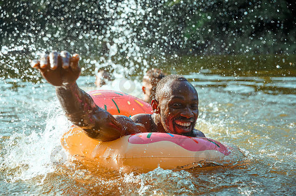 快乐的男人在河里嬉笑和游泳时玩得很开心。阳光明媚的河畔，快乐的男模特拿着橡胶圈做甜甜圈。夏日、友谊、度假、周末概念。