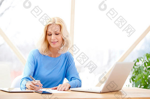 一幅金发碧眼<strong>的</strong>女商人坐<strong>在家</strong>里<strong>的办公</strong>桌旁，一边写东西，一边用笔记本电脑<strong>工作的</strong>照片。