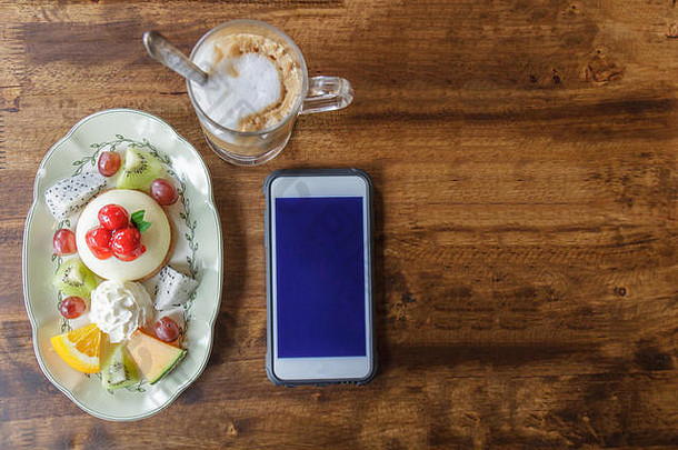 白盘子里的水果芝士蛋糕，咖啡和智能手机放在<strong>餐厅</strong>的木桌上。带黑屏的智能手机，可在sm上添加文字或其他内容
