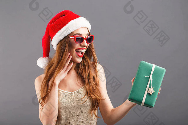 戴着红色圣诞老人帽子和红唇太阳镜的快乐迷人的女人对绿色包装的礼物感到惊讶，她笑着站在灰色背景上，新年、圣诞节、假期、纪念品、礼物、购物、折扣、商店、雪人圣诞老人、化妆、发型、狂欢节。