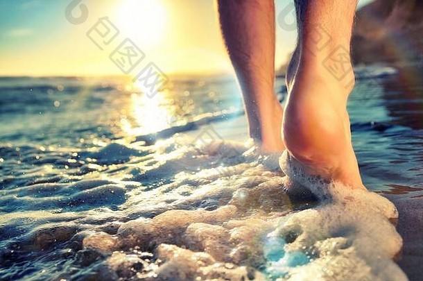 日落时分，一名男子赤脚在海滩上行走，在阳光和漂亮的镜头光晕下触水的特写镜头