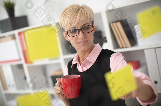 一个年轻女孩站在办公室里一块透明的贴纸板旁，手里拿着一个红色的杯子。