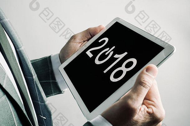 特写镜头中，一位身穿优雅灰色西装的白人年轻男子手里拿着一块数字平板电脑，屏幕上显示的数字是2018年，因为新年即将到来