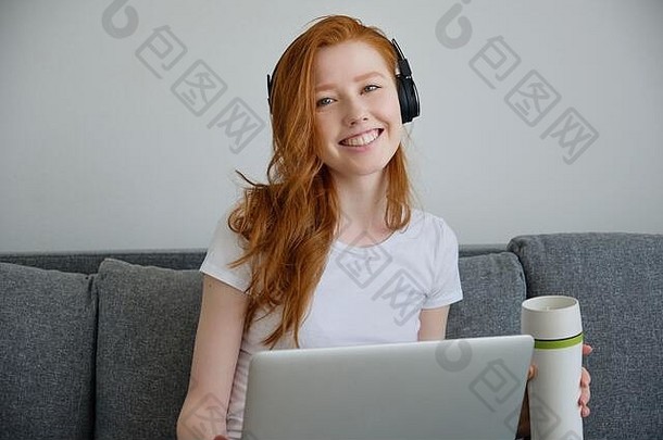 一个戴着耳机的可爱红发女孩对着放在膝上的笔记本电脑的摄像机微笑。